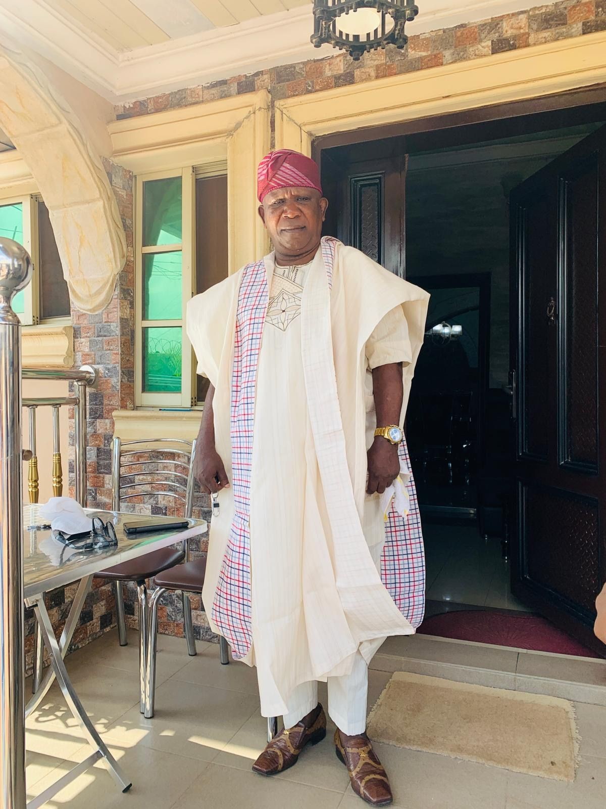 Man wearing traditional attire standing in front of a house with a brick wall and arched entrance.