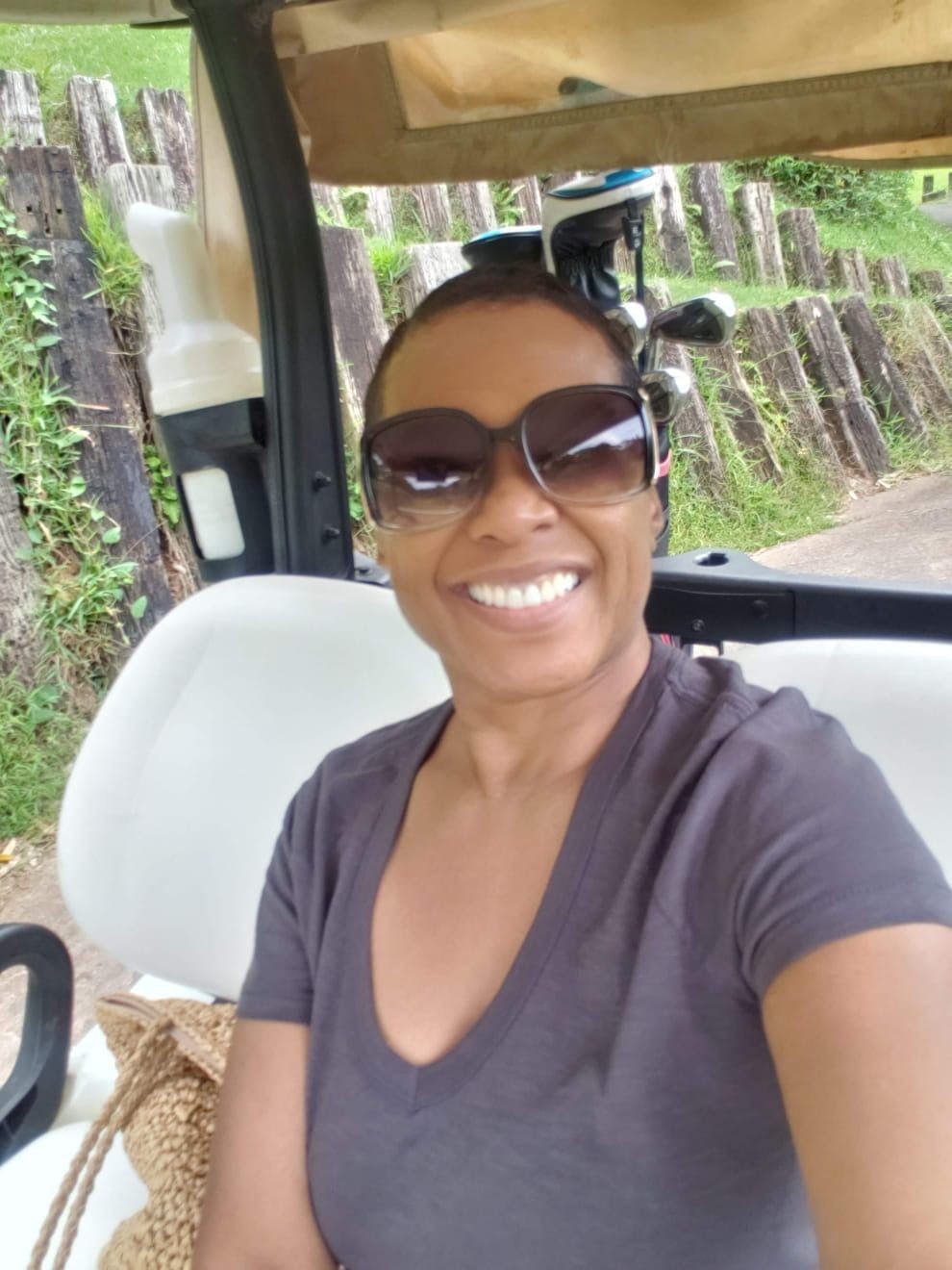 Person smiling and wearing sunglasses, sitting in a golf cart with golf clubs visible in the background.