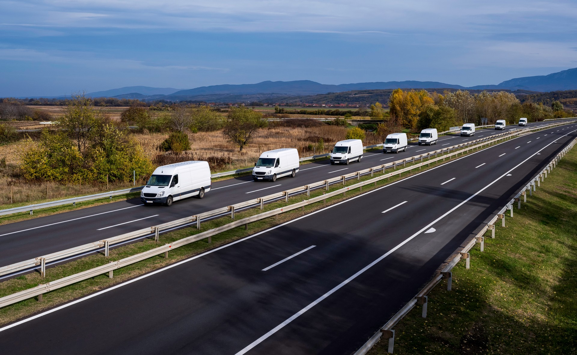 Eight white minivans are moving in a convoy on the highway. White delivery van on the highway. White modern delivery small shipment cargo courier van moving fast on motorway road to city urban suburb.